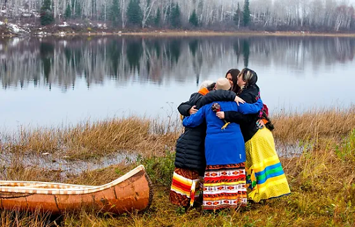 A group of women wearing ribbon skirts have a group hug next to a birchbark canoe by the river.